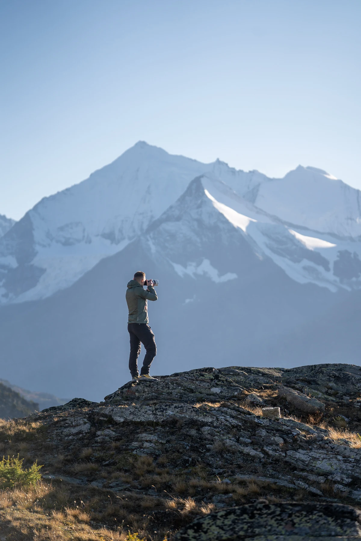 Kevin Buess fotografiert mit Kamera auf felsigem Untergrund vor schneebedecktem Walliser Alpengipfel, Schweiz