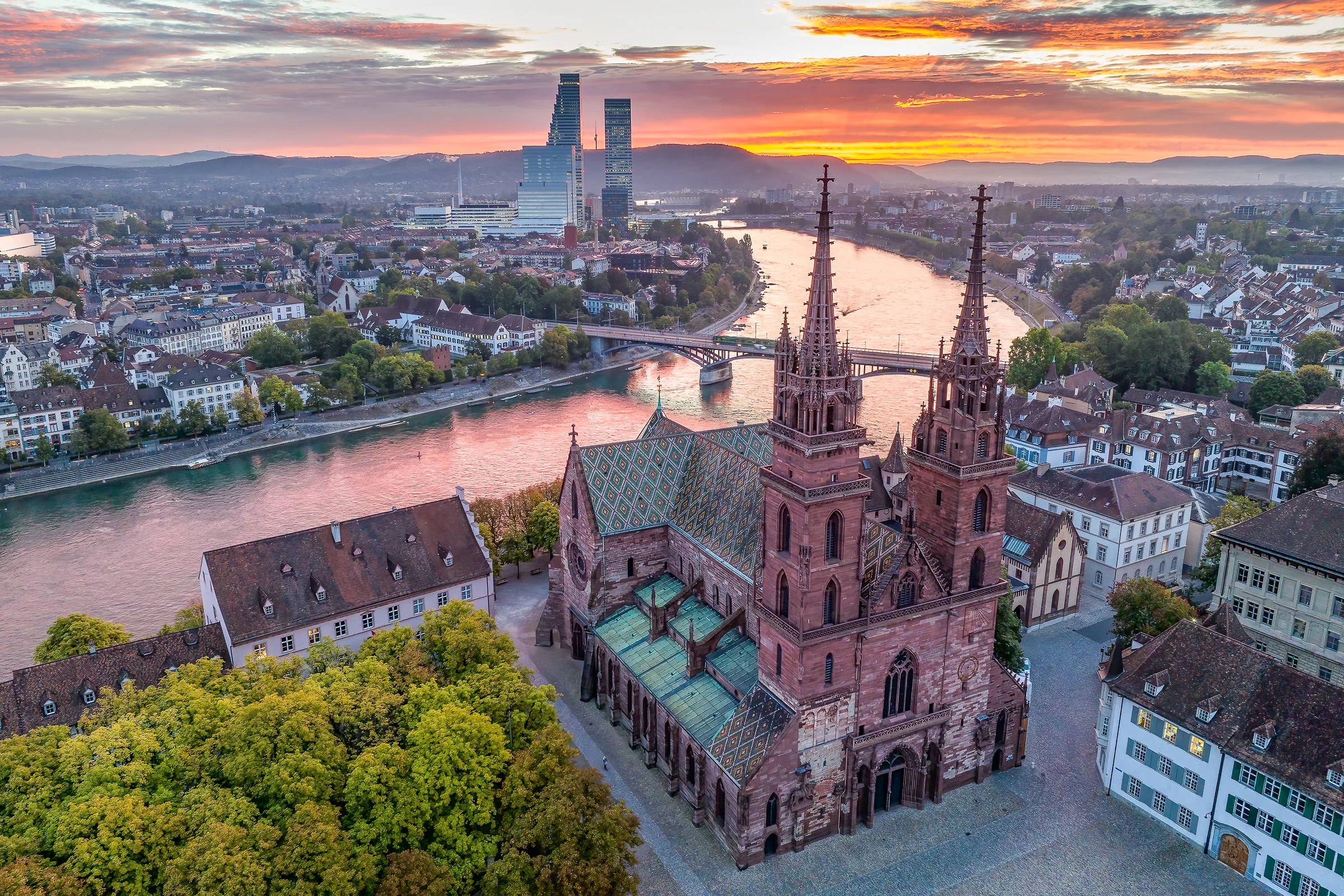 Luftaufnahme Basel Münster mit buntem Ziegeldach bei Sonnenuntergang, Rhein und Altstadt im Hintergrund