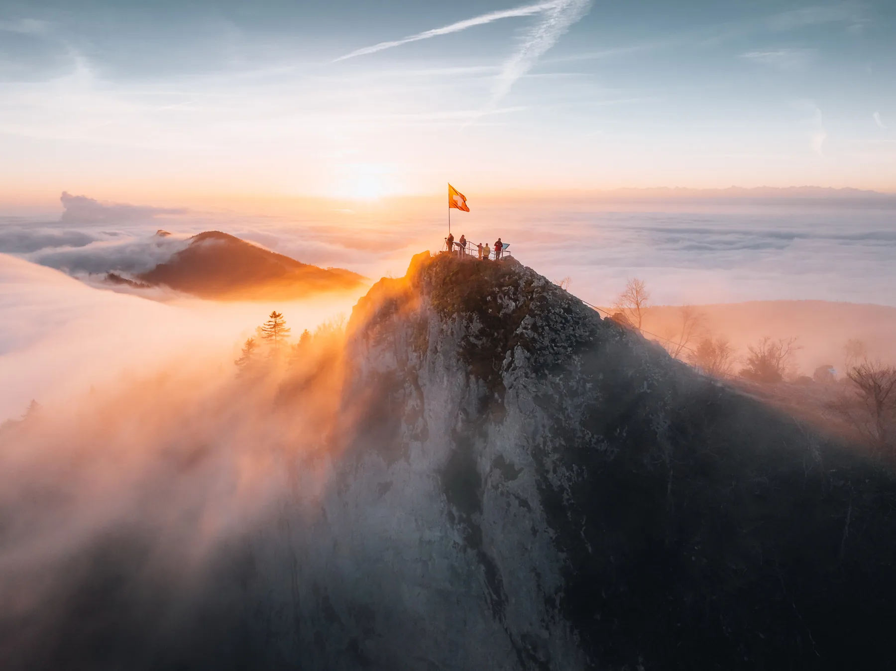 Felsgipfel mit Wanderern und Schweizer Flagge bei Sonnenaufgang über dichtem Nebelmeer, Schweizer Alpen, Belchenflue Baselland