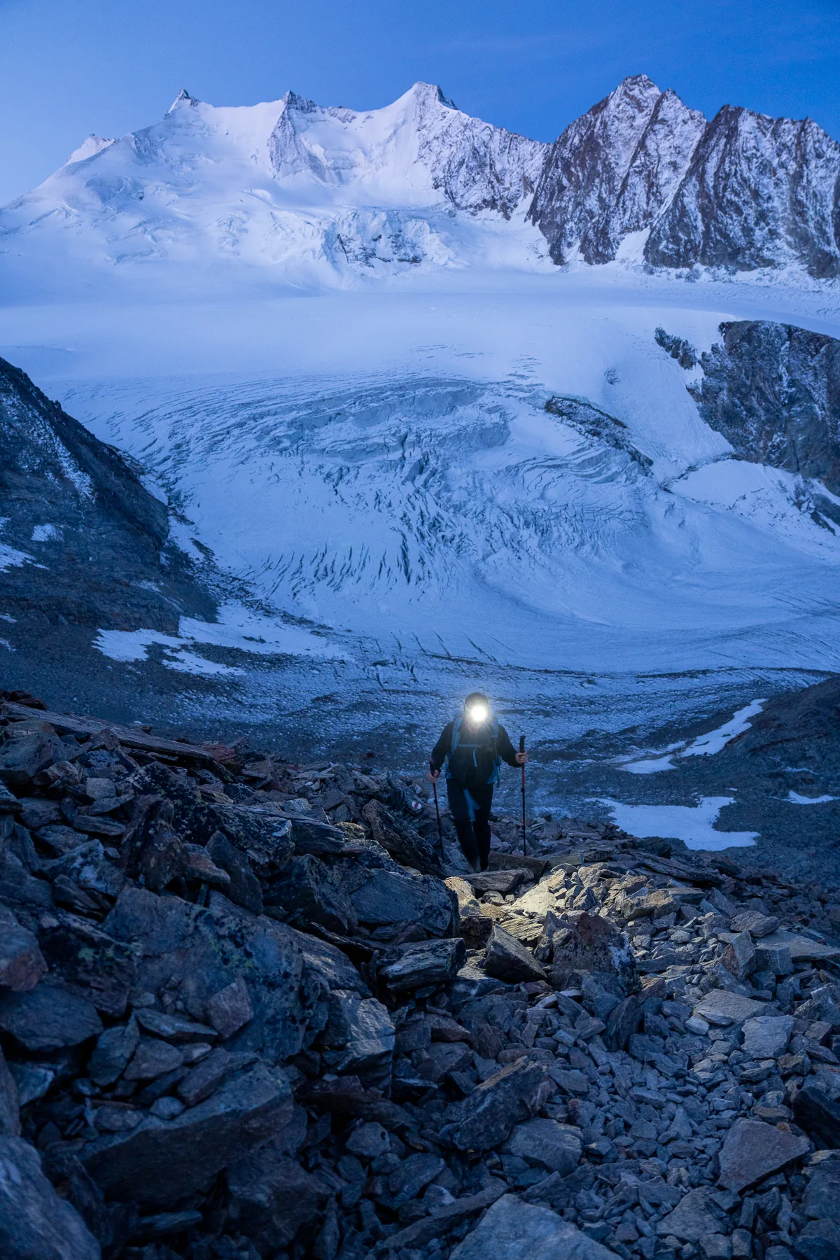Bergsteiger mit Stirnlampe klettert im Morgengrauen über Geröll, dahinter großer Gletscher und schneebedeckte Gipfel der Riedgletscher, Wallis, Schweiz