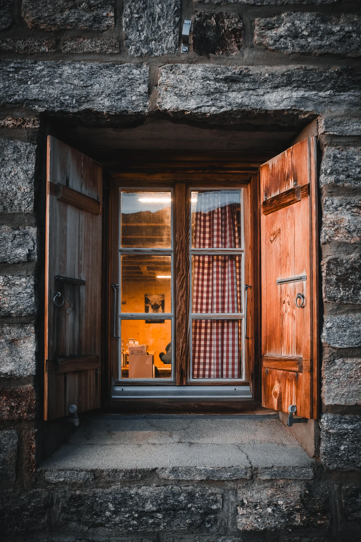 Geöffnete Holzfensterläden in rustikaler Steinmauer mit rot-weiss kariertem Vorhang und warmem Licht innen, Bordierhütte SAC, Wallis, Schweiz