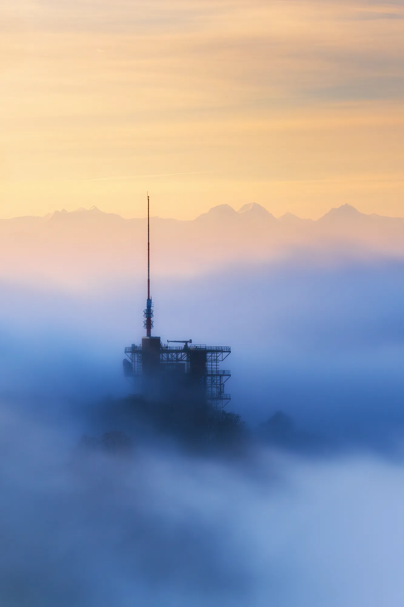 Sendeturm ragt aus dichtem Nebelmeer bei Sonnenaufgang, Alpen-Silhouette im Hintergrund, Schweiz, Wisen