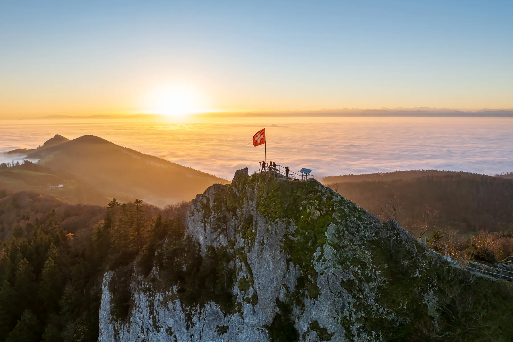 Schweizer Flagge auf felsigem Gipfel bei Sonnenaufgang, Wanderer auf Aussichtsplattform über Nebelmeer im Belchenflue, Baselland